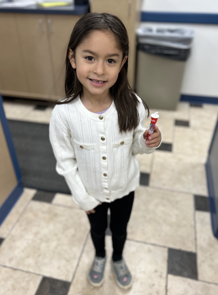 A young girl with long brown hair stands smiling in a room with tiled floors, wearing a white button-down cardigan and black leggings. She is holding up a small, red-and-white toy designed to look like a miniature soda cup.