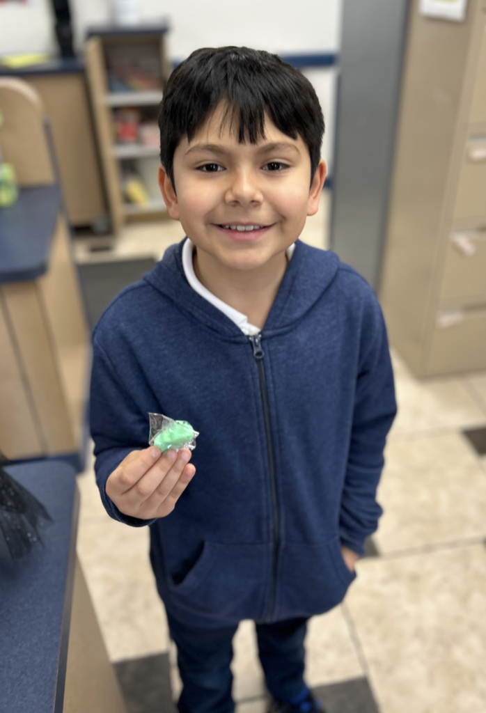 A smiling boy with dark hair, wearing a blue zip-up hoodie over a white shirt, stands indoors holding a small green item wrapped in clear plastic.