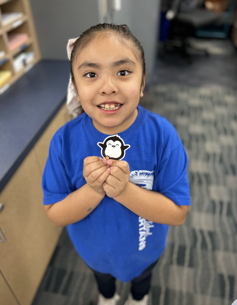 A young girl with dark hair pulled back into a ponytail and a white bow smiles brightly while holding a small cartoon penguin sticker with both hands. She is wearing a blue t-shirt with "SPARTANS" printed on the side and is standing in a classroom or office setting with a blurred bookshelf and patterned carpet in the background.