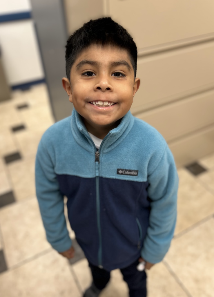 A young boy with short, dark hair smiles warmly at the camera. He is wearing a two-tone blue and navy fleece zip-up jacket. The photo is taken from a slightly high angle in a room with a tiled floor and a neutral-colored wall in the background.