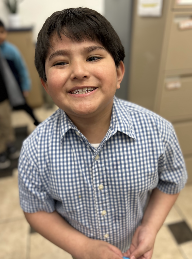 A young boy with short dark hair and a joyful smile stands in a room, wearing a blue and white checkered short-sleeve button-down shirt. The background is softly blurred, showing a tiled floor and a beige filing cabinet.