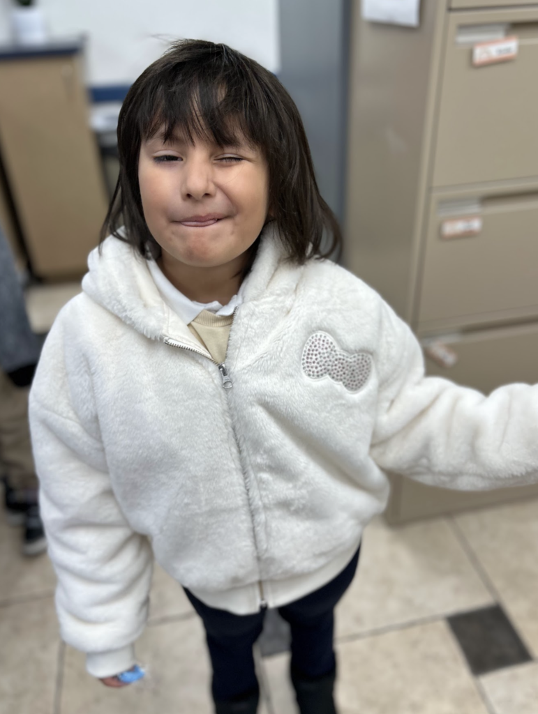 A young girl with dark hair makes a playful, winking face while wearing a white, fuzzy zip-up hoodie featuring a sparkly bow detail on the chest. She is standing in a room with a filing cabinet and tiled flooring visible in the background.