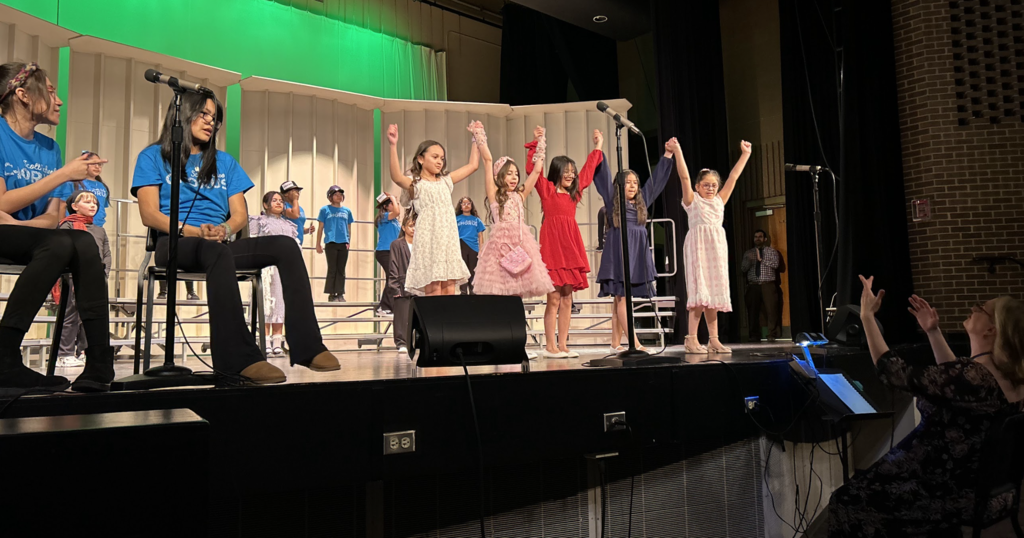 An on-stage performance featuring a diverse group of children at a school recital or chorus event. In the center, five young girls in colorful dresses stand in a row, smiling and holding their hands high in a triumphant "bow" pose. To the left, several other students in blue chorus T-shirts are seated or standing on risers. In the bottom right foreground, a music director with blonde hair is seen from behind, gesturing with both hands to lead the performance. The stage is equipped with professional microphones and monitors, set against a backdrop of white acoustic panels and a vibrant green screen.