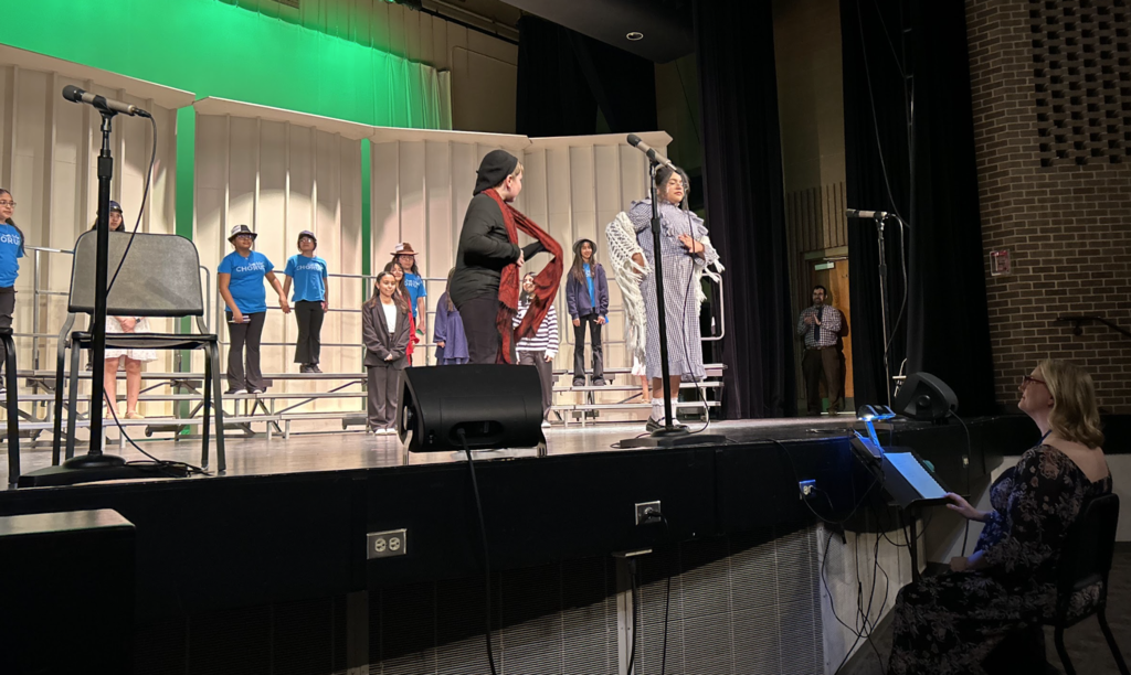 A wide shot of a school stage during a performance where a group of students are arranged on choral risers. In the foreground, two students in costume—one in a black outfit with a red scarf and another in a blue checkered dress with a white shawl—interact near a microphone. A woman sits at a music stand in the lower right corner, directing or accompanying the performance, while other students in blue "CHORUS" t-shirts stand behind them against a green-lit backdrop.