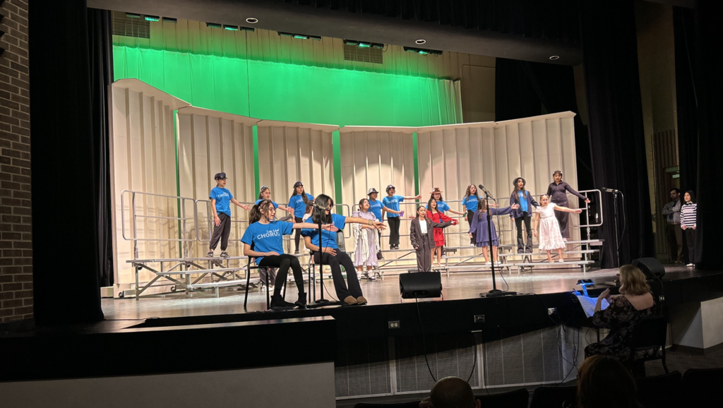 A wide shot of a student chorus performing on a theater stage. Several students stand on tiered risers, some wearing bright blue "CHORUS" t-shirts and others in formal dresses or suits, while two students sit in the foreground on chairs. They are all gesturing with their arms outstretched during a musical number. The stage features tall white acoustic shells illuminated by vibrant green overhead lighting, and a conductor is visible in the lower right corner guiding the performance from the orchestra pit.