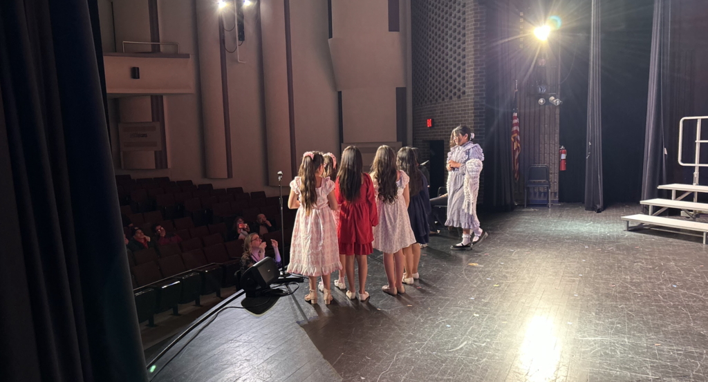 A group of young performers stands in a line on a theater stage, viewed from a side angle behind the curtain. Five girls with long hair are dressed in colorful dresses—pink, red, and floral patterns—facing away from the camera toward a student in a light purple dress who appears to be leading or speaking to them. The dimly lit auditorium seating is visible to the left, and a bright stage light creates a flare in the upper right, illuminating the dark wooden stage floor.