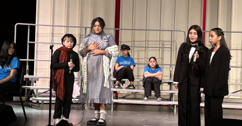 A medium-wide shot of 7 children in various costumes singing on a tiered stage, with several children sitting on metal risers in the background.