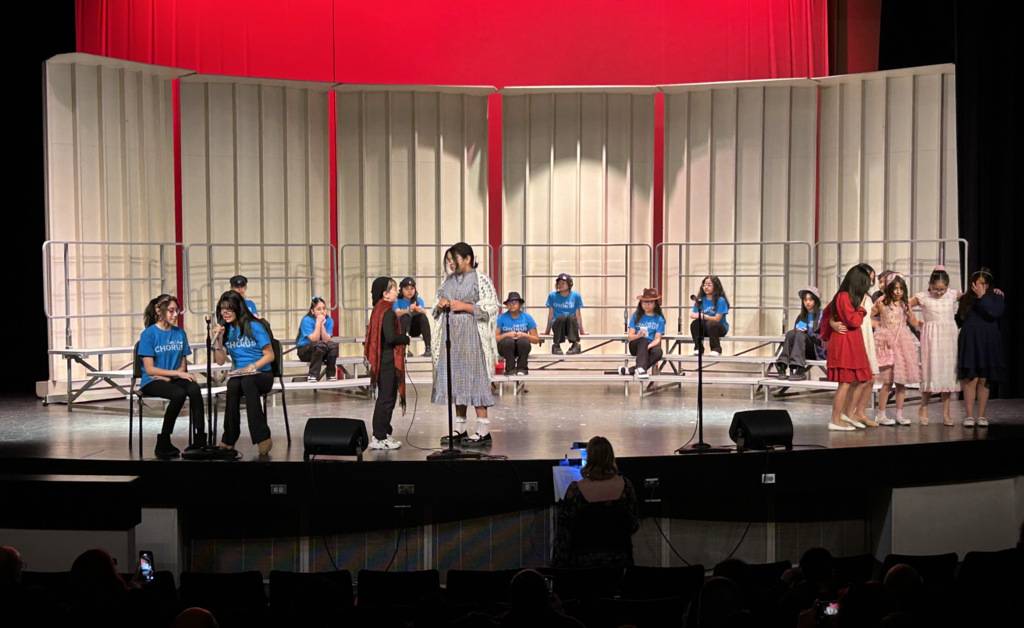 A wide shot of a school stage performance featuring a large group of students. In the center, two students in costume perform a scene with microphones, while others in blue "Chorus" t-shirts sit on tiered risers in the background. On the far right, a group of girls in colorful dresses stands together. The set features tall white acoustic panels against a vibrant red backdrop, with stage monitors and an audience visible in the foreground.