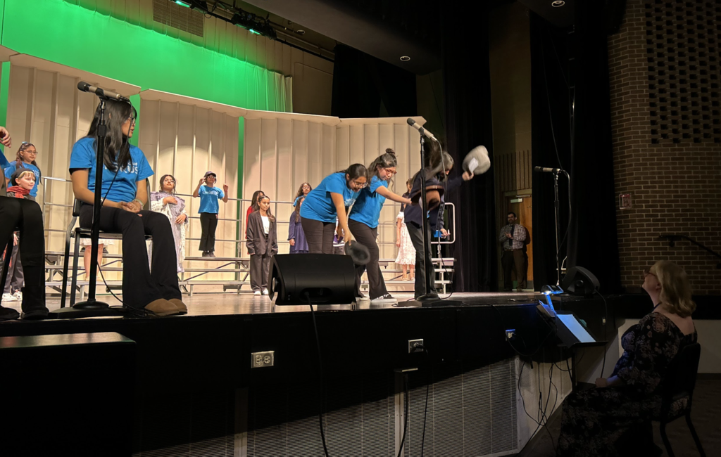 A group of students performing a musical number on a stage with a bright green backdrop. In the center, several students in blue T-shirts lean forward in a synchronized "hats off" gesture, holding hats toward the floor. To the left, a student sits in a chair near a microphone, while others stand on choral risers in the background. In the bottom right foreground, a music conductor with blonde hair sits in the audience area, looking up at the stage. The scene is captured from a low angle, highlighting the stage equipment and the energy of the school performance.