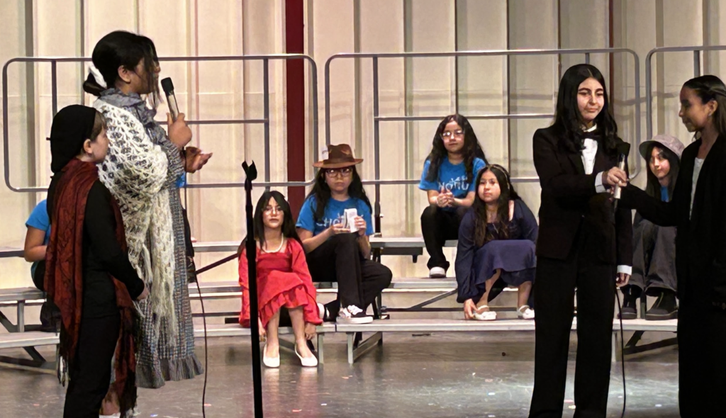 A medium-long shot captures a diverse group of seven young people performing a theatrical performance on a stage with risers. Four girls stand in the front of the stage, including one with a white shawl over her shoulders holding a microphone. Three of the children are sitting on metal risers behind the standing performers, including a person on the far left wearing a red ruffeled dress and a person wearing a brown cowboy hat. They stand against a background of white panels framed in red.
