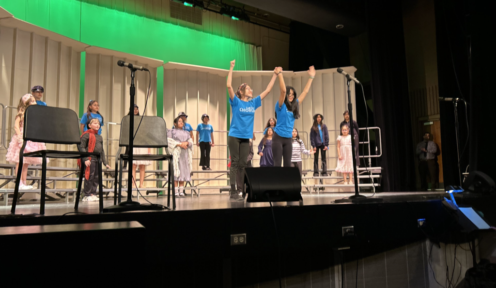 Two students in matching blue "CHORUS" t-shirts stand center stage with their arms raised and hands joined in a celebratory gesture. Behind them, other students in costumes and chorus shirts are positioned on tiered risers against white acoustic shells and a bright green backlit curtain. Microphones and stage monitors are visible in the foreground of the theater.