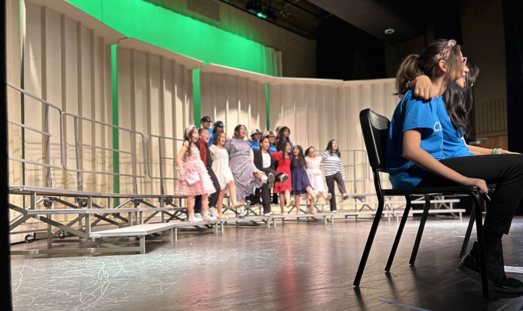 A group of diverse children are performing on a theater stage, standing on tiered risers against a backdrop of light-colored acoustic panels illuminated with green light. Most of the children are smiling and singing, dressed in a variety of colorful semi-formal outfits and casual school attire. In the foreground on the right, a girl in a bright blue t-shirt sits on a black chair, looking toward the group. The stage floor is dark and reflective, showing slight scuff marks from use.