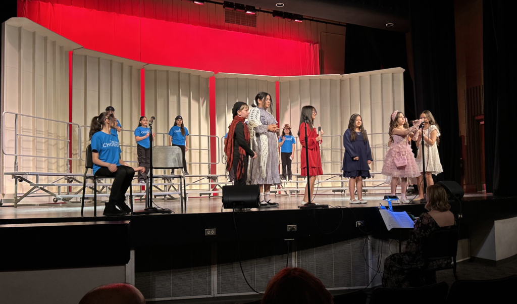 A group of students performs on a stage in front of white acoustic shells and a bright red backdrop. Several students stand in a line at the front of the stage wearing various outfits, including a red dress, a blue dress, and a pink ruffled dress, while others in blue "CHORUS" t-shirts stand on tiered risers behind them. A person sits at a piano in the lower right foreground, facing the performers.