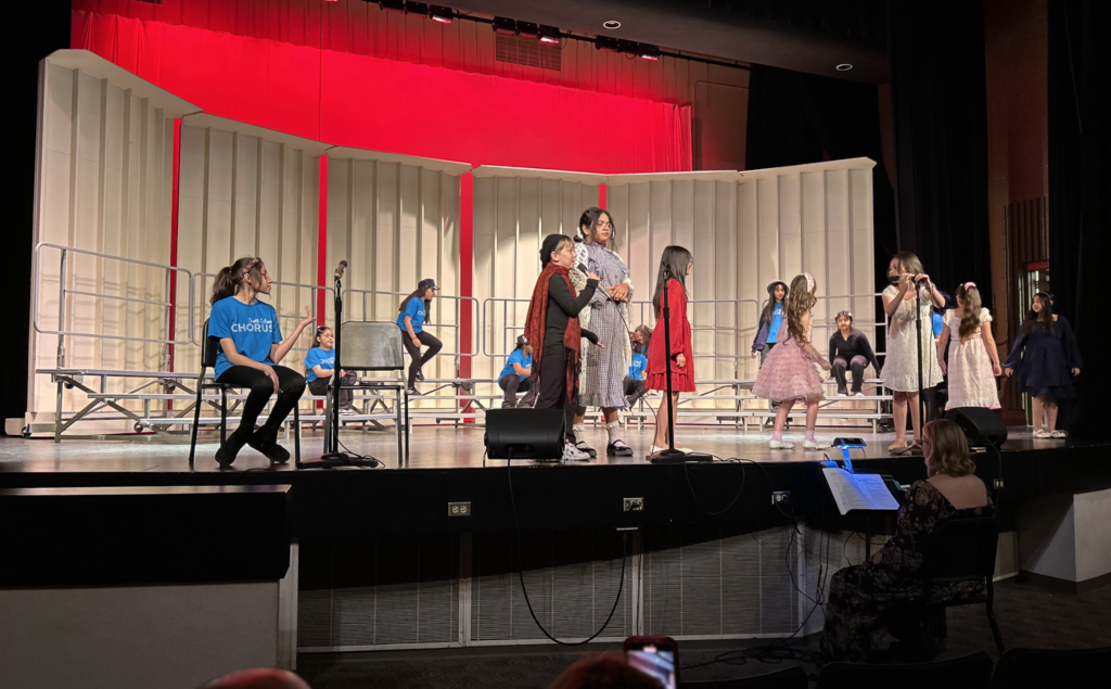 A group of middle-school-aged girls performs on a stage during a school production. Several girls in the center are dressed in costumes, including a red dress, a pink tutu, and a long plaid skirt, while others in the background wear blue t-shirts labeled "CHORUS." The stage features white acoustic shells, red backlighting, and tiered risers. In the foreground, an adult sits at a piano or music stand, accompanying the performance.