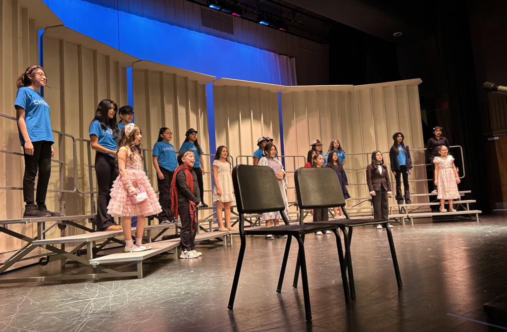A group of students stands on tiered risers on a stage during a performance. Some students wear bright blue t-shirts that say "CHORUS," while others are dressed in formal dresses, suits, or hats. Two empty black chairs sit in the foreground, and the background features white acoustic panels under blue stage lighting.