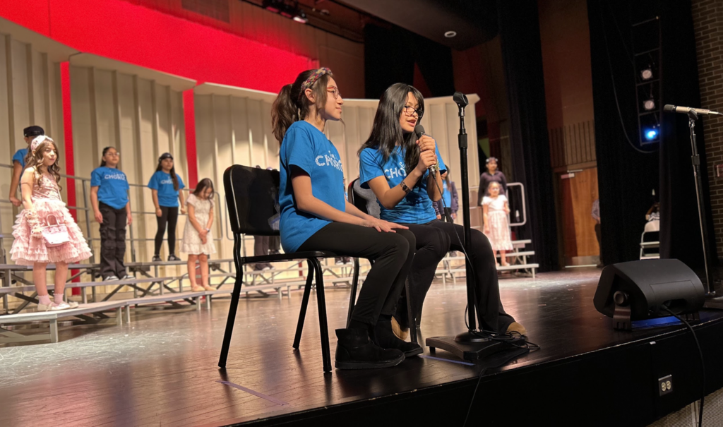 On a dimly lit stage, two young girls sit on chairs and speak into a microphone. Both wear blue "CHORUS" t-shirts and glasses. In the background, other students are on risers in a variety of outfits, including blue t-shirts and formal wear, under a backdrop with red accents.