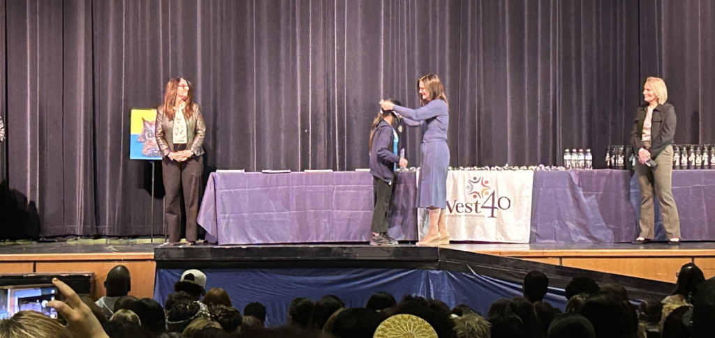 On a stage with dark curtains, a woman in a blue dress presents an award to a student in front of a table draped in purple. To the left, another woman stands by a colorful painting on an easel, while a third woman stands to the right near a row of water bottles. An audience is visible in the foreground, and a banner on the table reads "West40."