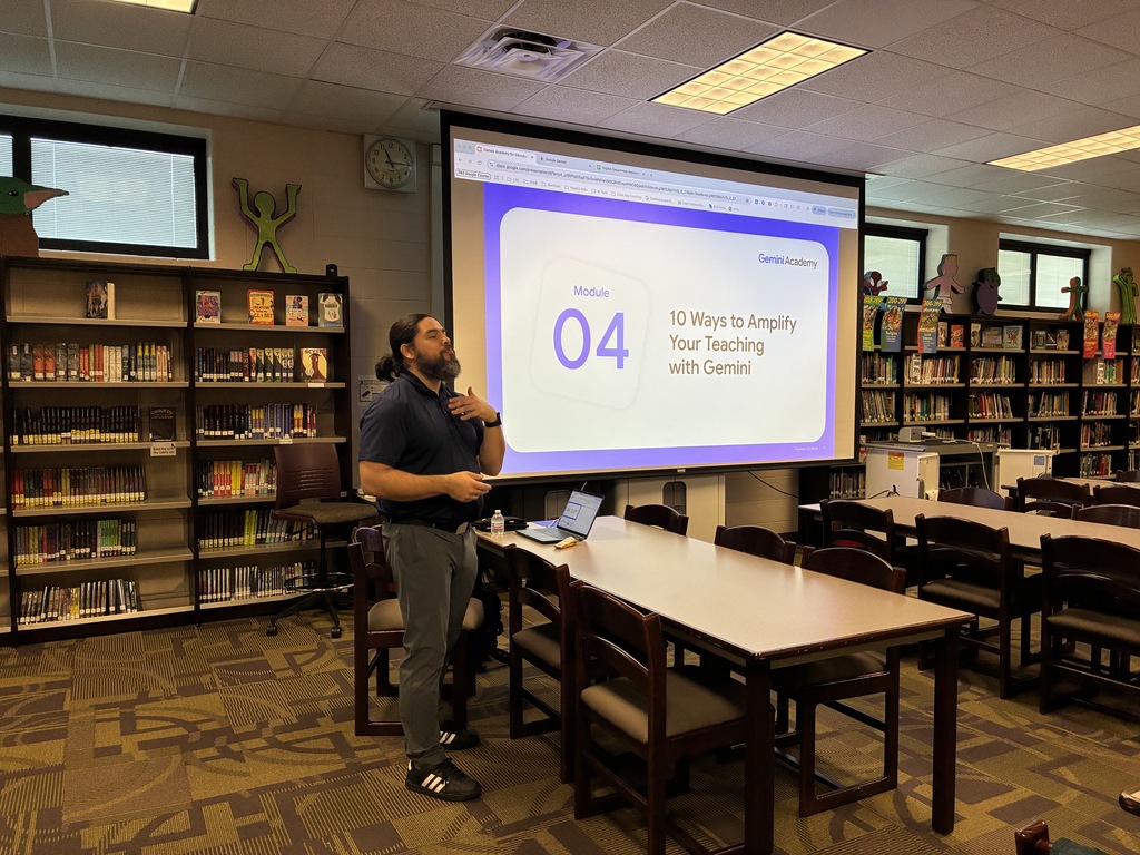 An educator with long dark hair tied back and a beard stands in a well-lit school library, gesturing with one hand toward his chest while presenting. He is positioned next to a long table with a laptop, facing a large projection screen displaying a "Gemini Academy" slide titled "Module 04: 10 Ways to Amplify Your Teaching with Gemini." The background features organized bookshelves filled with books, student-oriented decorations, and large windows, creating an engaging academic atmosphere.