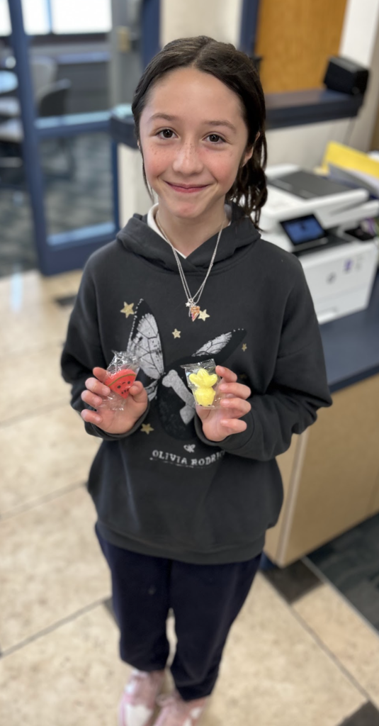 A smiling girl with dark hair stands in an office-like setting, wearing a dark grey hoodie with a butterfly graphic. She is holding up two small, wrapped erasers: one shaped like a red watermelon slice and the other like a yellow pineapple. The background shows a printer and tiled flooring.