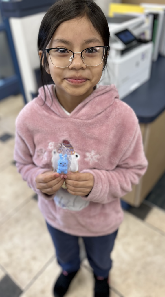 A young girl with dark hair and glasses smiles warmly while standing in what appears to be an office or school setting. She is wearing a soft, fuzzy pink hoodie featuring a polar bear graphic and is holding a small, bright blue bunny figurine in both hands. The background is slightly blurred, showing a printer and tiled flooring.