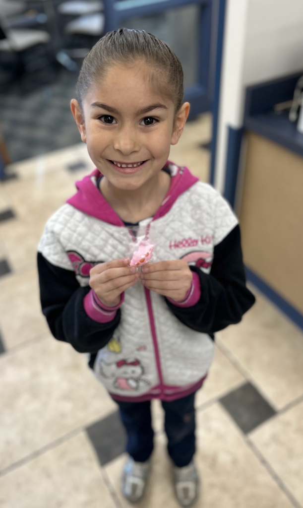 A high-angle, full-length shot shows a young girl with her hair pulled back into a neat bun, smiling warmly at the camera. She is wearing a white, black, and pink Hello Kitty zip-up hoodie over dark pants and grey shoes. She holds a small, pink, heart-shaped object—possibly a piece of candy or a small craft—between her fingers at chest level. The background features a beige and black checkered tile floor and the blurred interior of a room with blue accents. The lighting is bright and clear, giving the photograph a cheerful, candid feel.