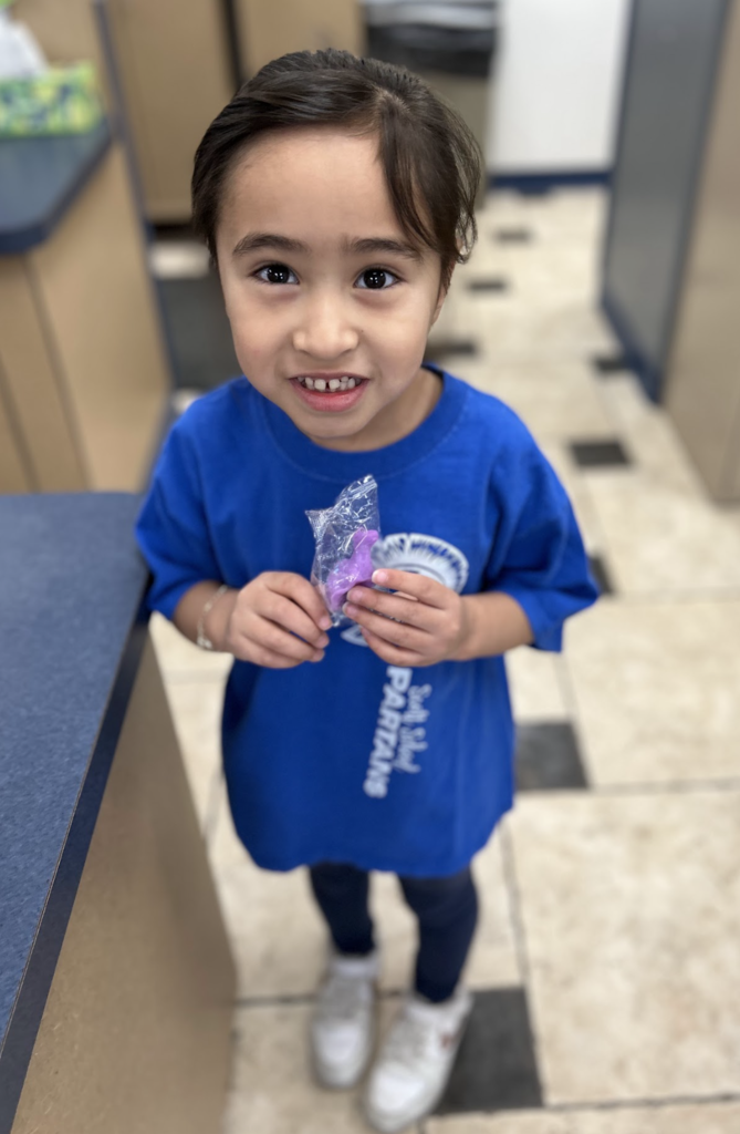 A young child with dark hair and a wide, gap-toothed smile stands in a room with tiled floors. They are wearing a bright blue t-shirt that says "PARTANS" and dark leggings, holding a small, purple, dinosaur-shaped object inside a clear plastic wrapper.