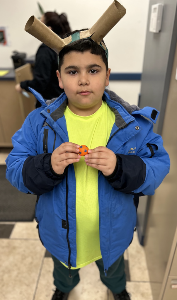 A medium, full-length shot shows a young boy standing in an indoor setting, wearing a bright blue winter jacket over a lime green t-shirt and dark green pants. On his head, he wears a homemade headband made of green paper and two cardboard tubes taped to the sides to resemble horns or ears. He is holding a small orange ball with blue dots between his hands at chest level, looking directly at the camera with a neutral expression. The background is slightly blurred, showing a person with dark hair in a black sweater, a beige tiled floor, and office-like furniture. The lighting is bright and even. The style of the image is a photograph.