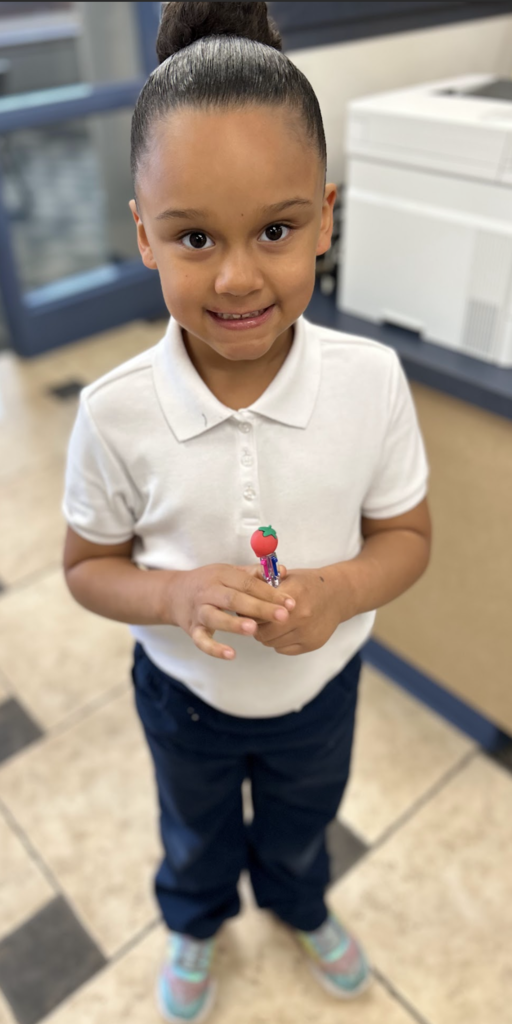 A young girl with her hair styled in a high bun smiles at the camera while standing in a room with tiled floors. She is dressed in a white polo shirt and dark trousers, holding a decorative pen topped with a small red strawberry. The background features a desk and a white office printer.