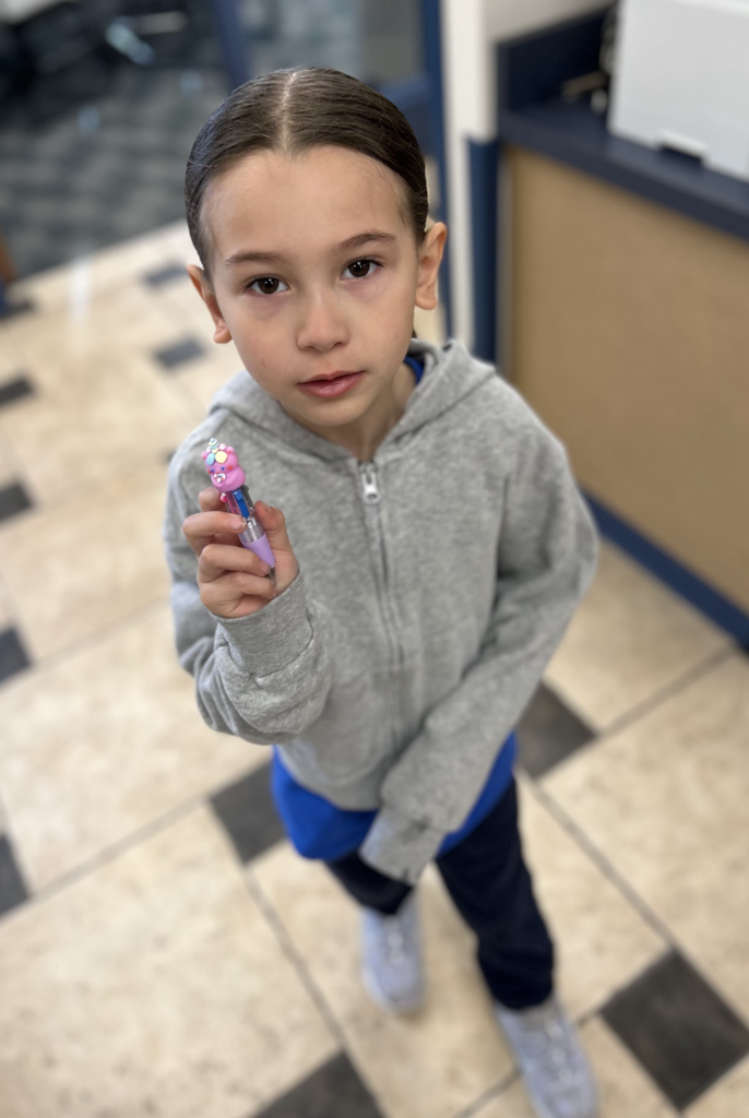 A young girl with dark hair pulled back looks toward the camera while holding up a multi-color clickable pen topped with a pink character. She is wearing a grey zip-up hoodie over a blue shirt and dark pants, standing on a checkered tile floor in an indoor setting.