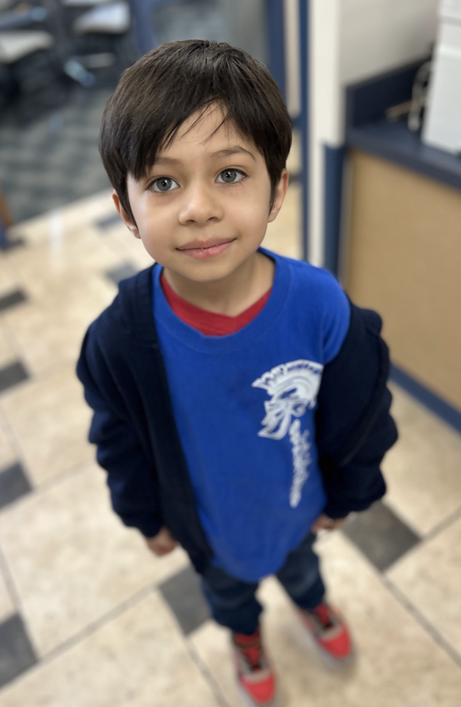 A portrait from above captures a young boy with dark hair and blue-green eyes wearing a blue T-shirt with a Spartan helmet logo over a red-collared shirt and an open black hoodie. He stands on a tiled floor with black and tan squares in an office environment, gazing directly up at the camera with a soft expression. The background elements include furniture and another person, slightly out of focus.