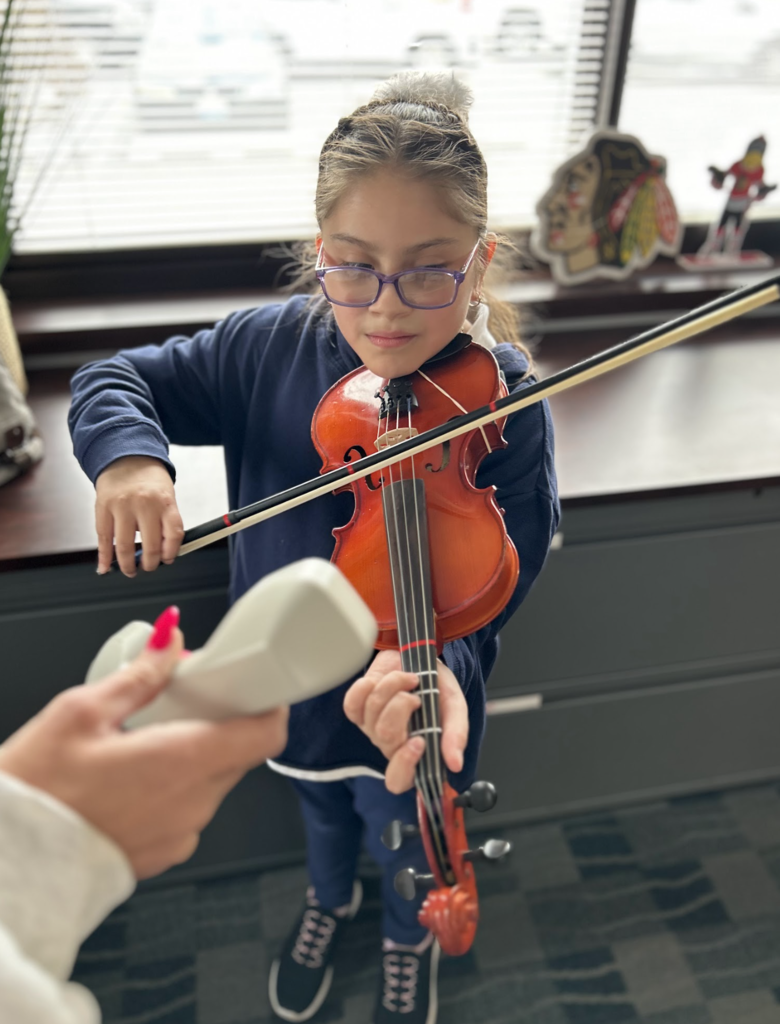 A medium shot shows a young person with light-brown braided hair pulled into a high ponytail and wearing purple-framed glasses, a dark blue pullover sweater, dark blue pants, and black and white lace-up sneakers looking down at a red violin in their hands. They are holding a bow in their left hand and placing it on the violin and their other hand is on the fingerboard of the violin as if ready to play. A hand with bright pink nail polish is visible in the bottom-left corner and is holding up a light-colored object to the person and the red violin. The background shows a dark desk and window with blinds. Behind the desk, an object resembling a feather headdress is visible and an object resembling a figure or hockey player. The person is standing on a dark grey patterned carpet. The light is natural and coming from the window. The style of the image is a photograph.