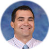 A headshot of a smiling man with short dark hair, wearing a pink and white checkered dress shirt and a light blue tie, set against a blurred blue background and framed within a circular crop.