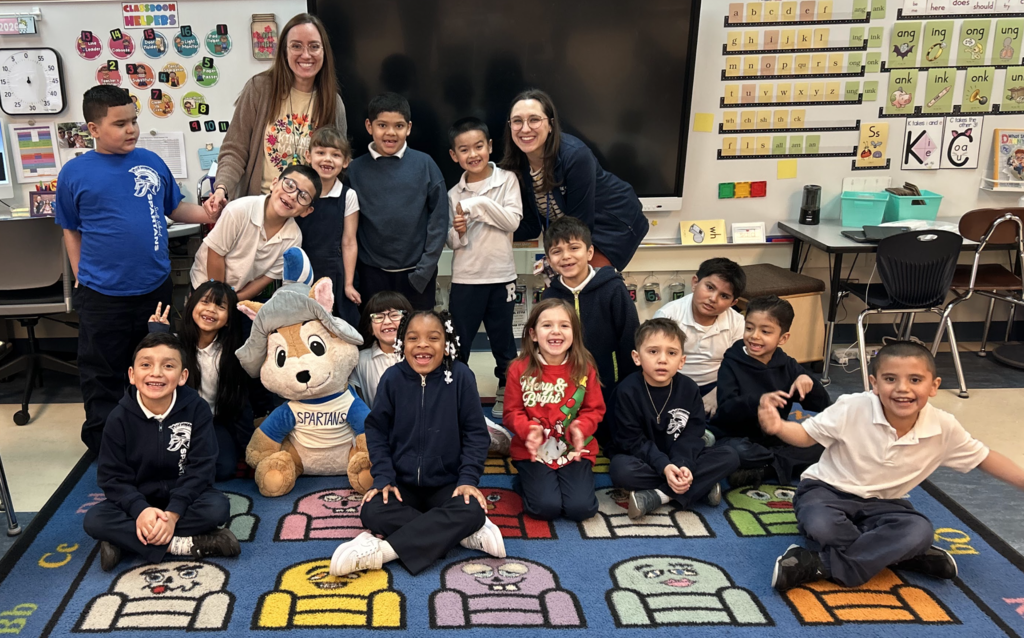A smiling female teacher stands with her 12 students and a life-sized mascot dressed as a SPARTANS football player in a primary school classroom, where they are grouped around a large blue area rug with colorful cartoon character faces. A large whiteboard and alphabet chart are on the back wall.