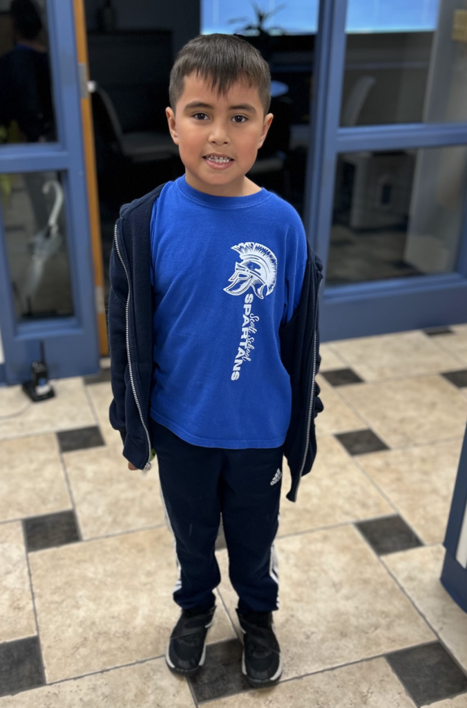 A young boy smiling and standing in a school hallway, wearing a blue t-shirt featuring the "Scott School Spartans" logo and mascot, a dark zip-up hoodie, and navy athletic pants.