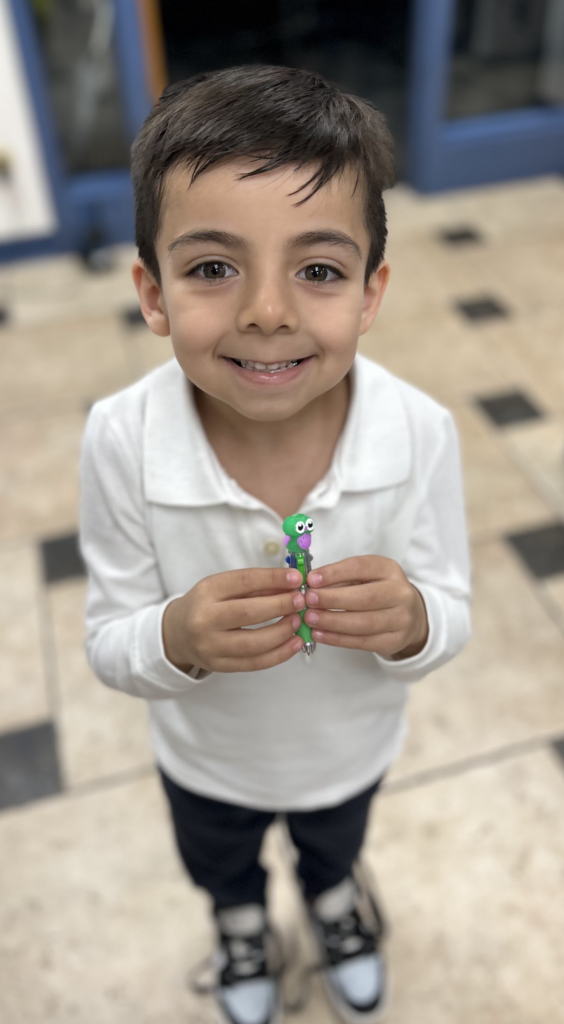 A young boy with dark, styled hair smiles broadly while holding a multi-colored pen with a green owl topper in both hands. He is wearing a white long-sleeve polo shirt and dark pants, standing on a light-colored checkered tile floor with dark accents.
