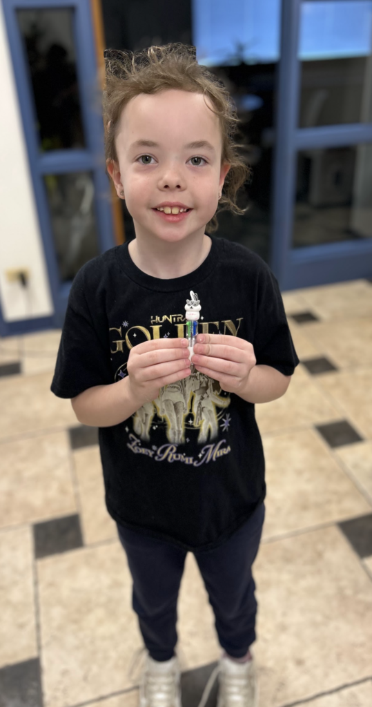A young girl with brown curly hair smiling and holding a small, white multi-color shuttle pen with a cartoon animal topper. She is wearing a black T-shirt featuring "Golden" text and three figures, dark blue pants, and white sneakers, standing on a checkered tile floor in front of a blue-framed doorway.