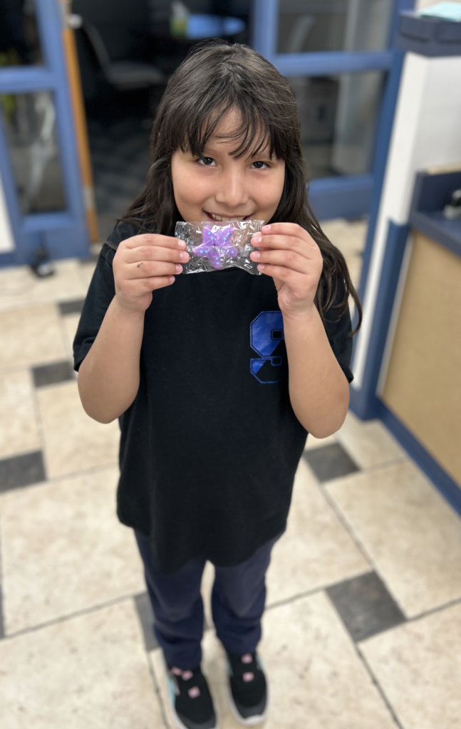 A smiling young girl with long brown hair holding up a small, purple starfish-shaped object in clear plastic packaging, wearing a black t-shirt and standing on a checkered tile floor.
