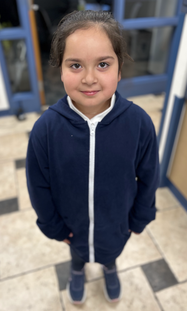 A young girl with brown hair and eyes, smiling and wearing a navy blue zip-up hoodie over a white shirt, standing on a tiled floor in a room with a blue door frame in the background.
