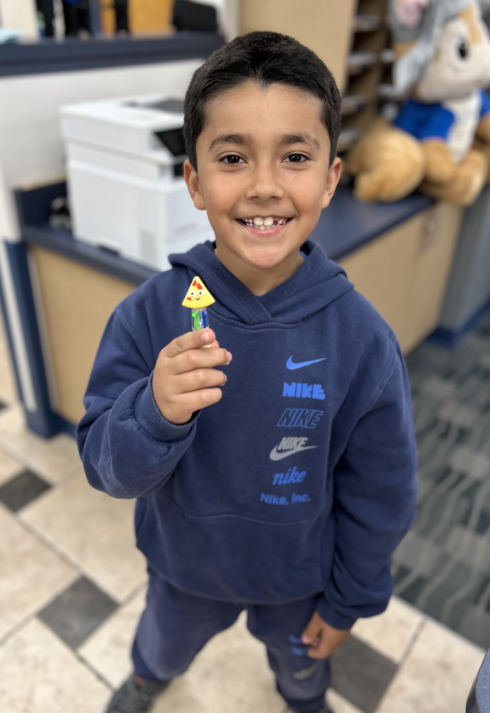A medium shot of a young boy smiling as he holds up a multi-colored pen with a cartoonish wedge-of-pizza charm on the top, in an office or school hallway with other people and a stuffed animal in the background. The boy wears a navy blue hooded sweatshirt that features a stack of Nike logos.