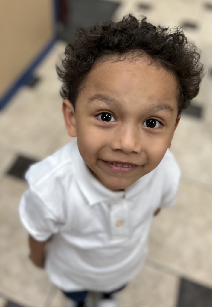 A high-angle close-up portrait of a young boy with curly dark hair and brown eyes looking up at the camera with a slight smile. He is wearing a white polo shirt and standing on a tan and black checkered tile floor.