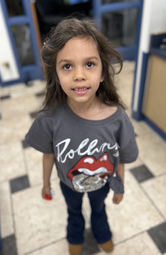 A young girl with long, wavy brown hair looking upward with a smile, wearing a gray Rolling Stones graphic t-shirt and dark pants, standing on a checkered tile floor.