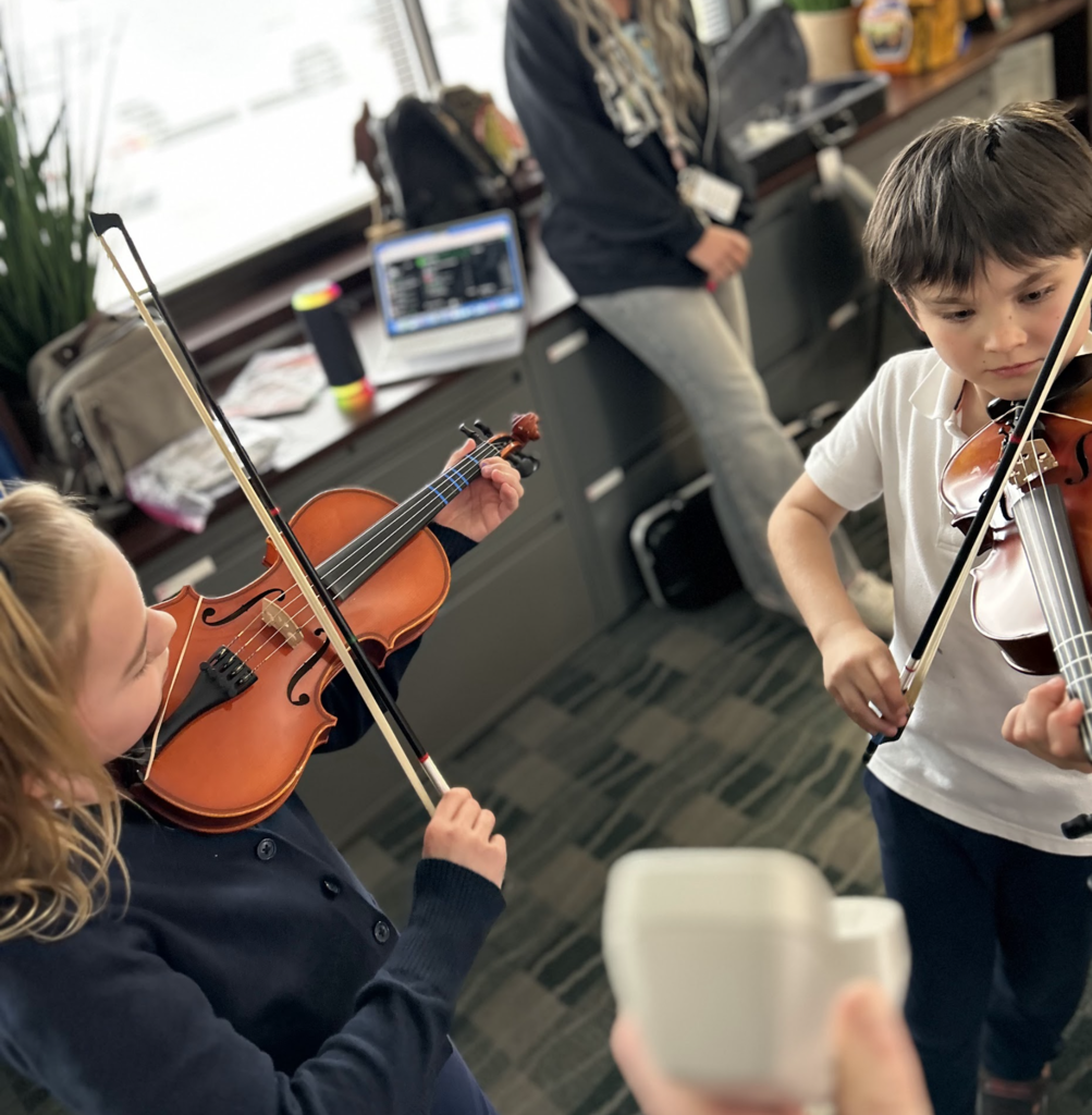 A medium-high angle photograph of two young students, one girl and one boy, actively playing violins during a lesson in a classroom, with another blurred adult figure and instruments in the background.
