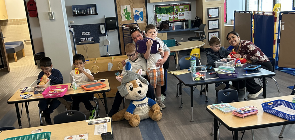 A wide-angle photograph of a busy early childhood classroom with multiple people engaged in activities. In the center-right, two female educators and three young boys are gathered around a blue table. One educator hugs a standing boy holding a purple tablet, while a seated boy looks on. Next to them, a second smiling educator sits at the table with a seated boy. On the floor in front of this group sits a large, plush wolf mascot wearing a blue and white "SPARTANS" jersey. To the left, two more boys are seated at their own small wooden table, one eating and the other looking down, with trays and an orange tablet. The classroom background features shelves, cubbies, bulletin boards, a water cooler, and an open bathroom doorway, all under warm indoor lighting.