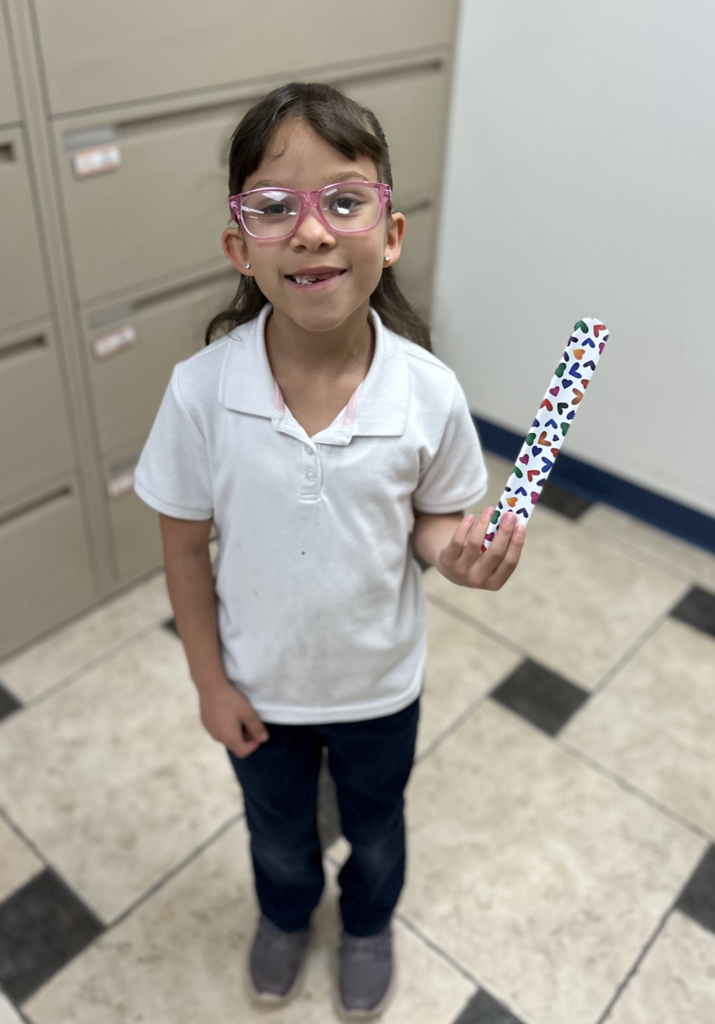 A young girl with glasses and a missing front tooth stands smiling, holding a colorful slap bracelet decorated with multi-colored hearts. She is wearing a white short-sleeed polo shirt, dark navy pants, and grey sneakers. The photo is taken from a high angle, showing a light-colored tiled floor and neutral filing cabinets in the background.