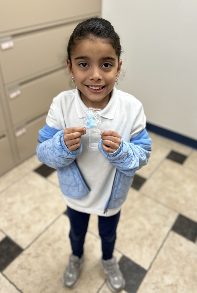 A young girl stands smiling in an office setting, holding up a small, clear plastic package containing a light blue toy. She is wearing a white collared polo shirt, a light blue quilted jacket, dark pants, and grey sneakers. The photo is taken from a high angle, showing a checkered tile floor and tan filing cabinets in the background.