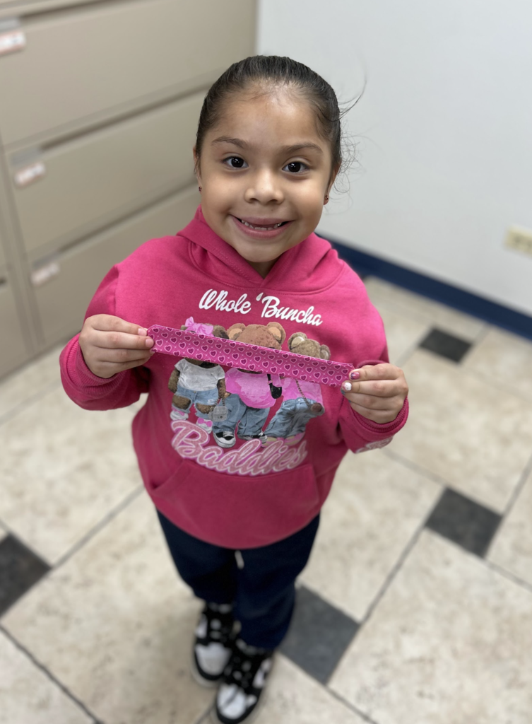 A young girl stands smiling in an office setting, holding up a pink headband patterned with small hearts. She is wearing a vibrant pink hoodie featuring a "Whole 'Buncha Baddies" bear graphic, dark pants, and black and white sneakers. The photo is taken from a high angle, showing a tiled floor and neutral-colored filing cabinets in the background.