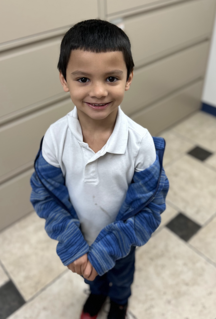 A young boy stands smiling in an office-style room, looking directly at the camera. He is wearing a white short-sleeved polo shirt and a blue striped zip-up hoodie that is resting slightly off his shoulders. The photo is taken from a high angle, showing a light-colored tiled floor with dark accents and tan filing cabinets in the background.