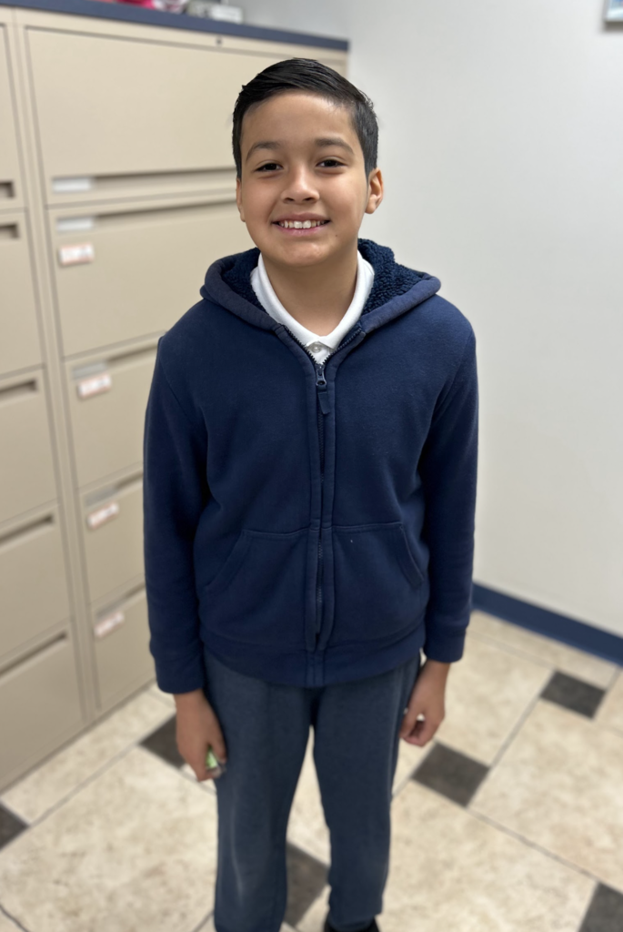 A young boy stands centered in a room, smiling directly at the camera. He is wearing a dark navy blue zip-up hoodie over a white collared shirt and matching dark blue trousers. The background features a neutral-colored wall, tan filing cabinets, and a light-colored tiled floor with a dark checkered pattern.