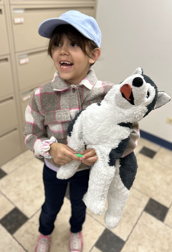 A young girl stands smiling and laughing while holding a large, plush husky stuffed animal. She is wearing a light blue baseball cap, a grey and pink plaid flannel jacket, dark pants, and pink sneakers. The husky toy is black and white with its tongue sticking out. The background shows a neutral-colored room with a tiled floor and tall filing cabinets.