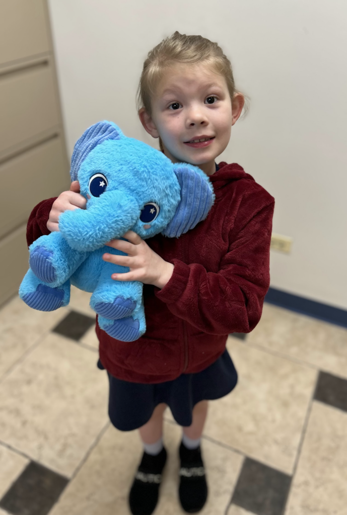 A young girl stands smiling while hugging a large, bright blue plush elephant. She is wearing a fuzzy maroon zip-up jacket over a dark navy skirt, paired with black slip-on shoes. The photo is taken from a high angle in an indoor setting with a tiled floor and a neutral-colored wall and filing cabinet in the background.
